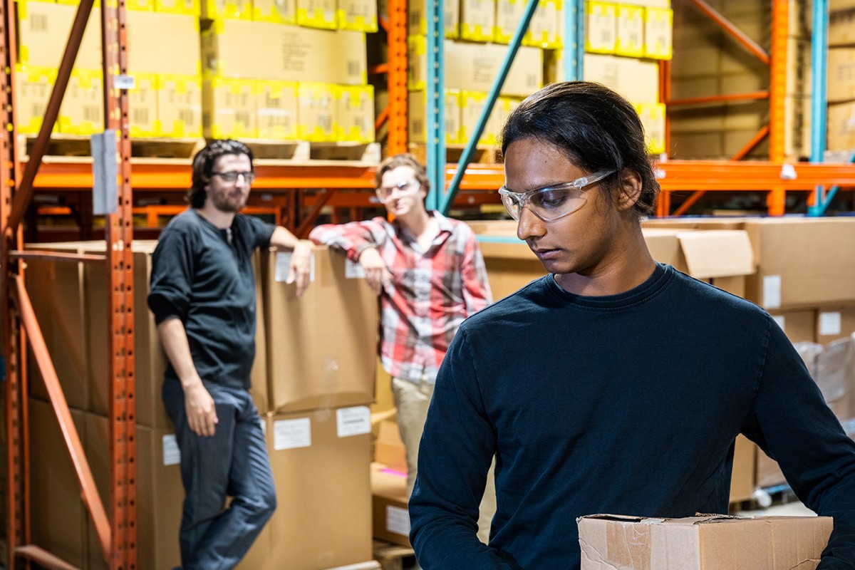 A warehouse worker looks down uncomfortably while two colleagues watch and whisper in the background, suggesting workplace bullying.