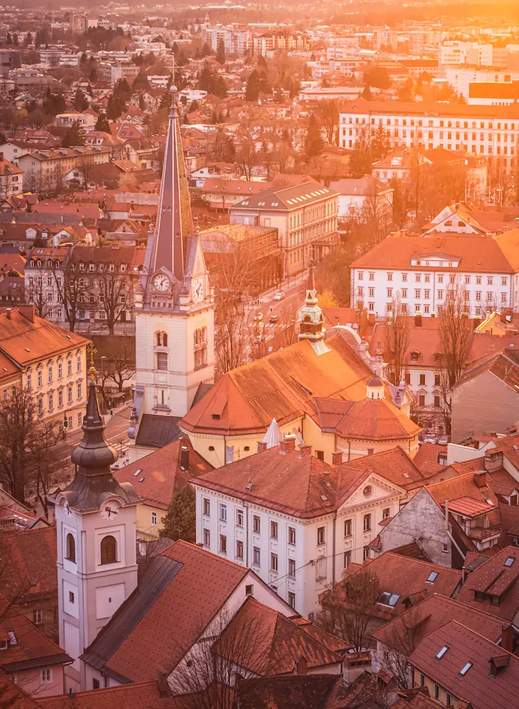 High-angle view of Ljubljana, Slovenia, at sunset, showing several historic church steeples rising above a sea of traditional orange-tiled roofs, all bathed in a warm golden glow.