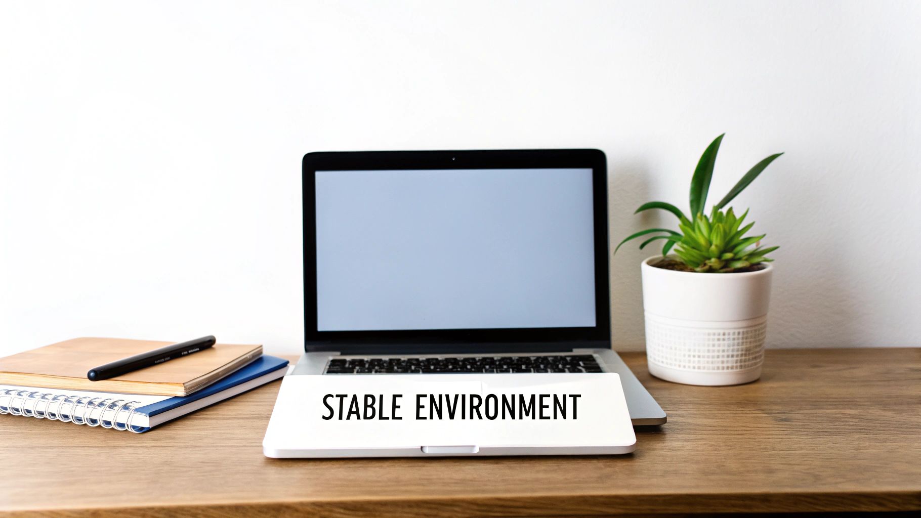 A laptop on a wooden desk with notebooks, a plant, and a 'Stable Environment' sign.