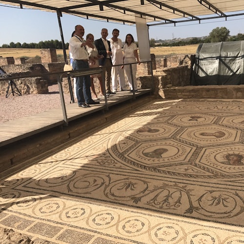 다섯 명의 사람들이 실외에 있는 큰 차양 아래 전시된 고대 모자이크 타일을 관찰하면서 보도를 걷고 있습니다.