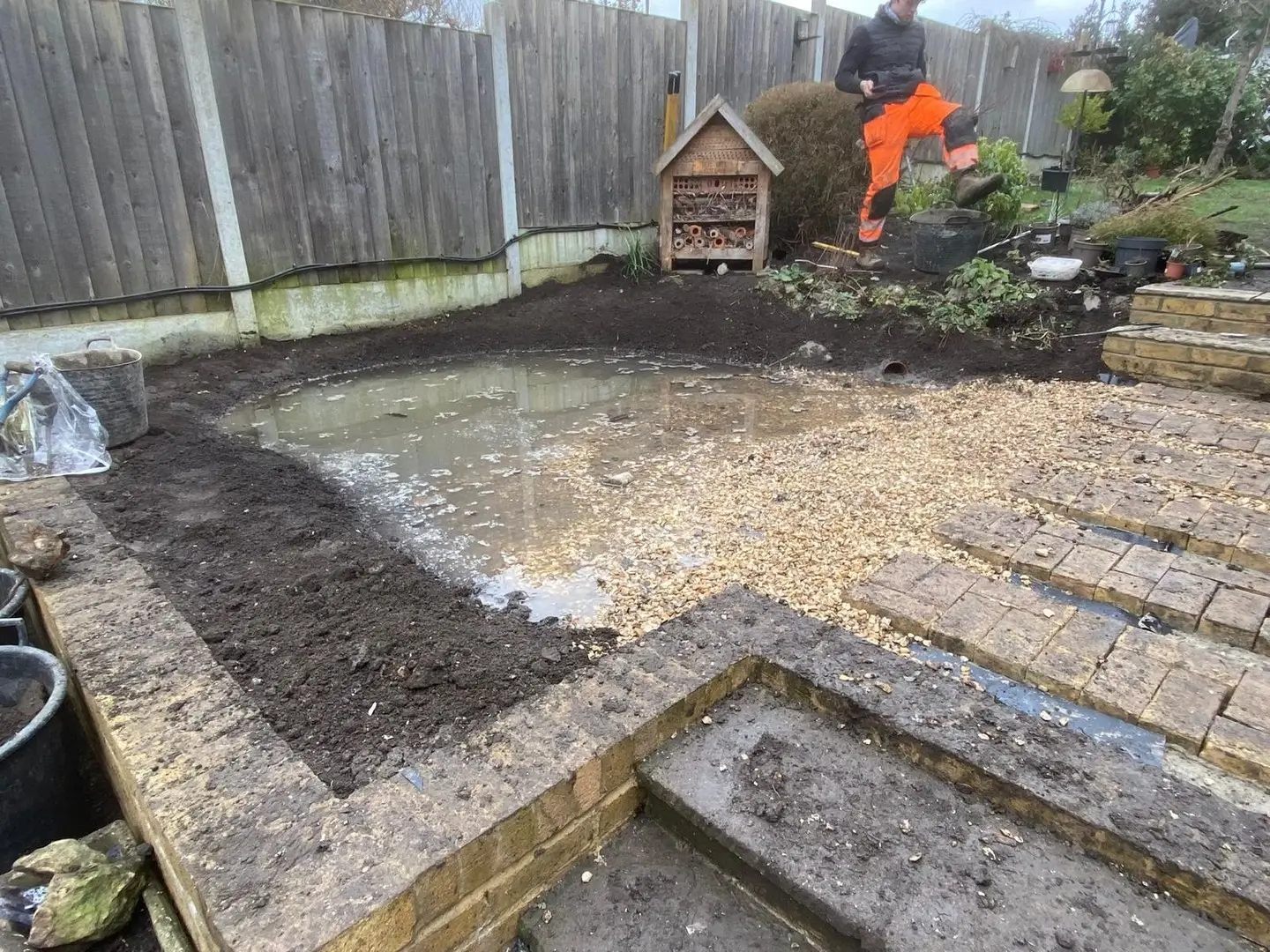 Construction area with freshly laid sand and gravel, a worker in an orange vest operates machinery in the background.