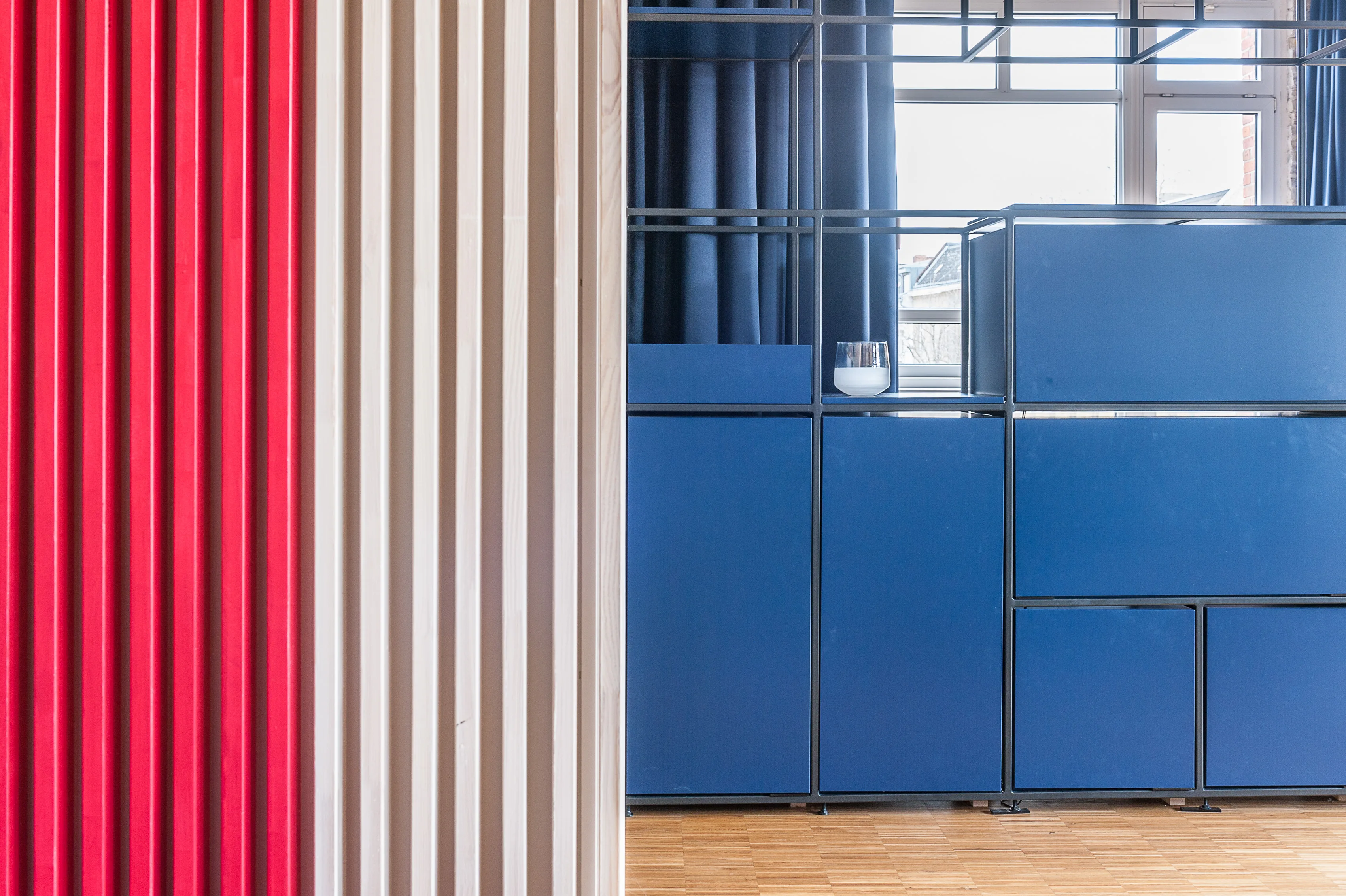 Detail at BASILICOM with a vertical slatted wall in red and beige stripes next to blue modular cabinets in a black steel frame, a navy curtain and a glass tumbler on the counter.