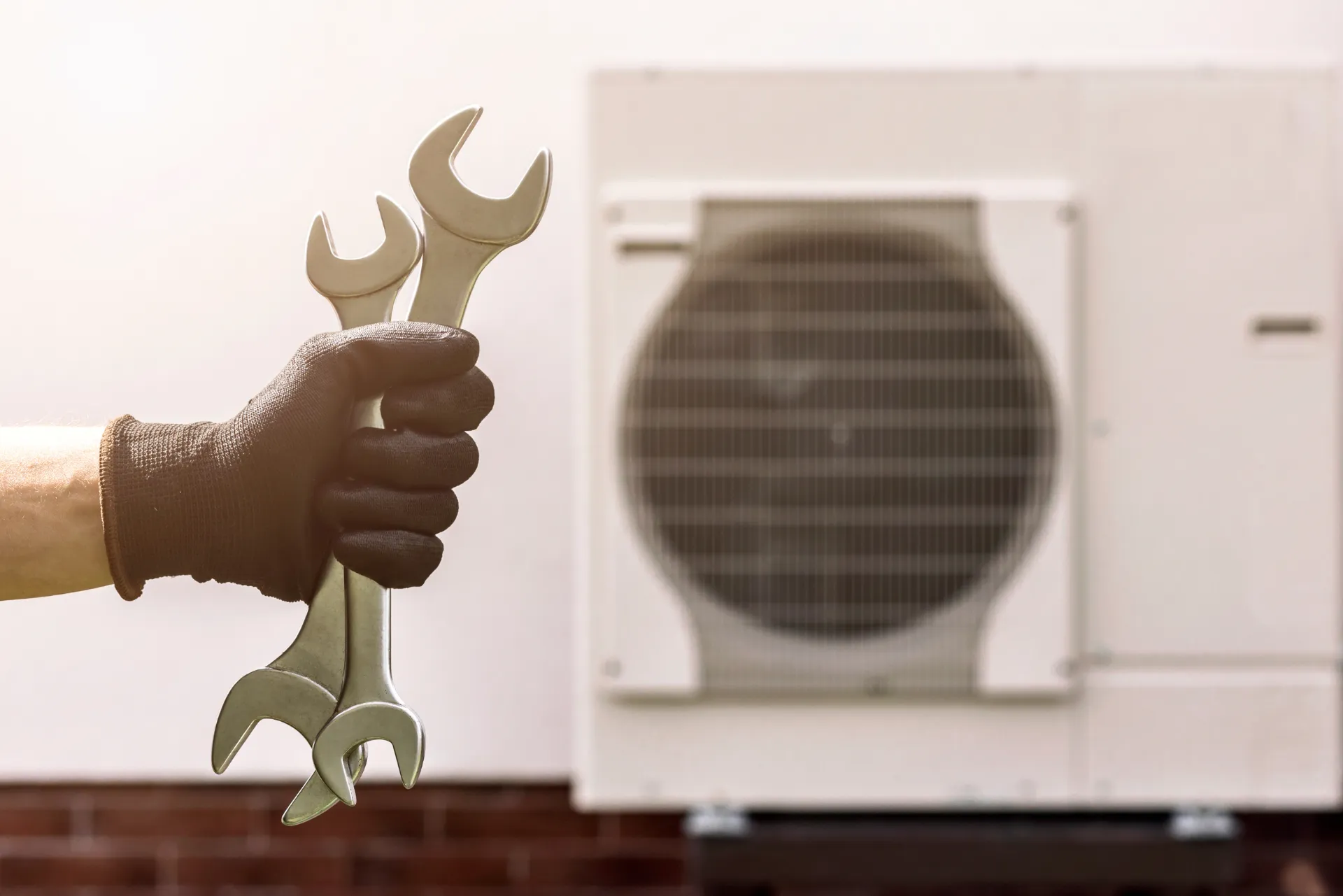 Die Hand eines Handwerkers welcher gerade zwei Schraubenschlüssel im Vordergrund hält. Im Hintergrund befindet sich eine Wärmepumpe, verschwommen, an der Hauswand montiert.