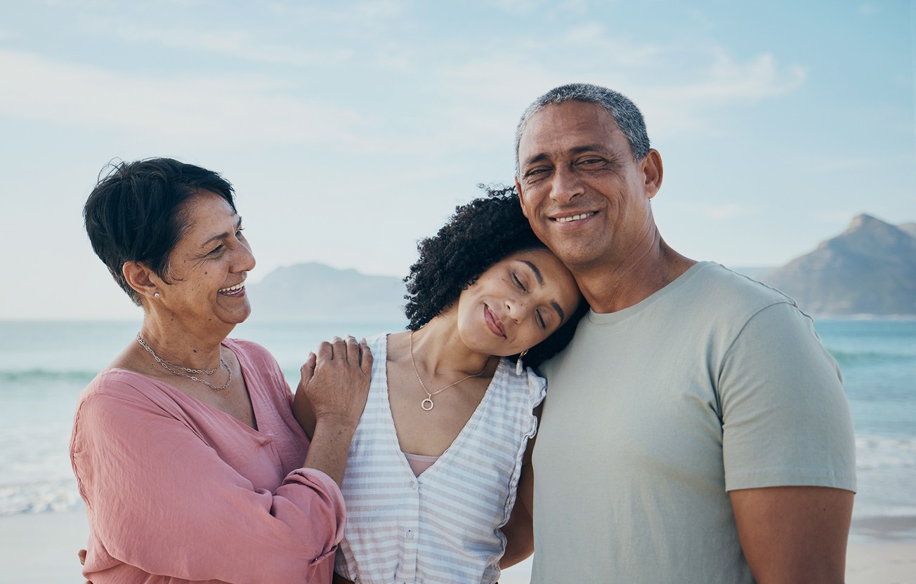 A smiling, diverse, multigenerational family of three—a mother, father, and adult daughter—embracing on a sunny beach