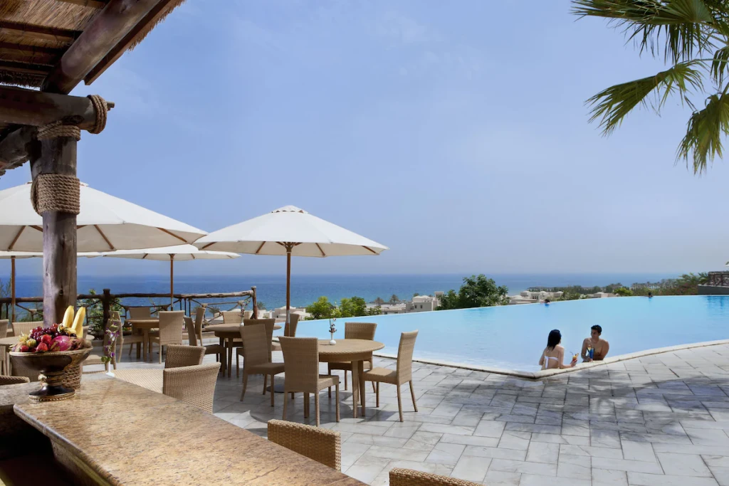 Scenic view of the Cove Rotana Resort's beach, featuring soft sand, clear water, and palm trees under a blue sky.