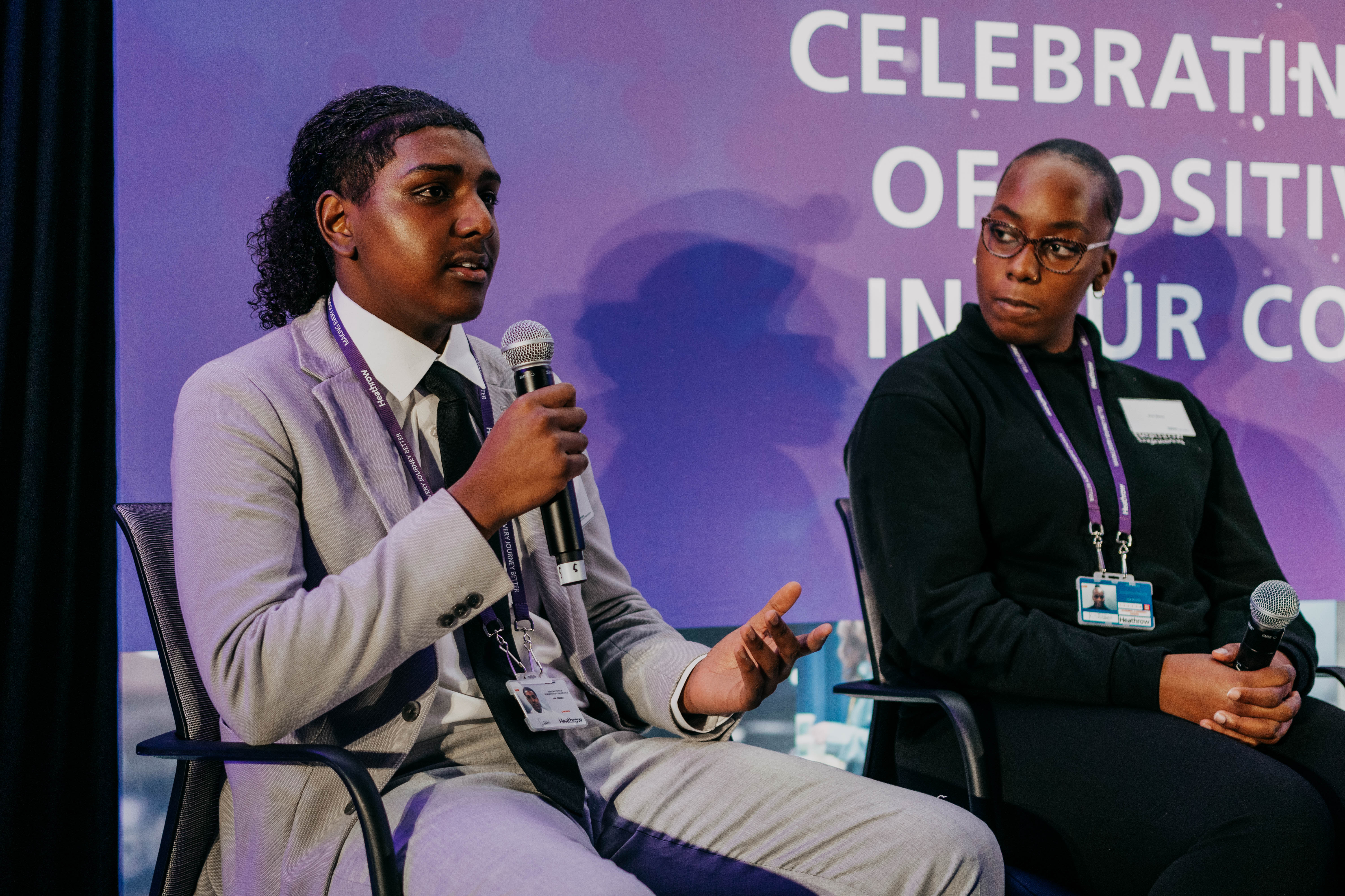 A young man in a suit speaks into a microphone while sitting on stage during a panel discussion. Beside him, a young woman listens attentively. They are in front of a large purple backdrop with white lettering.