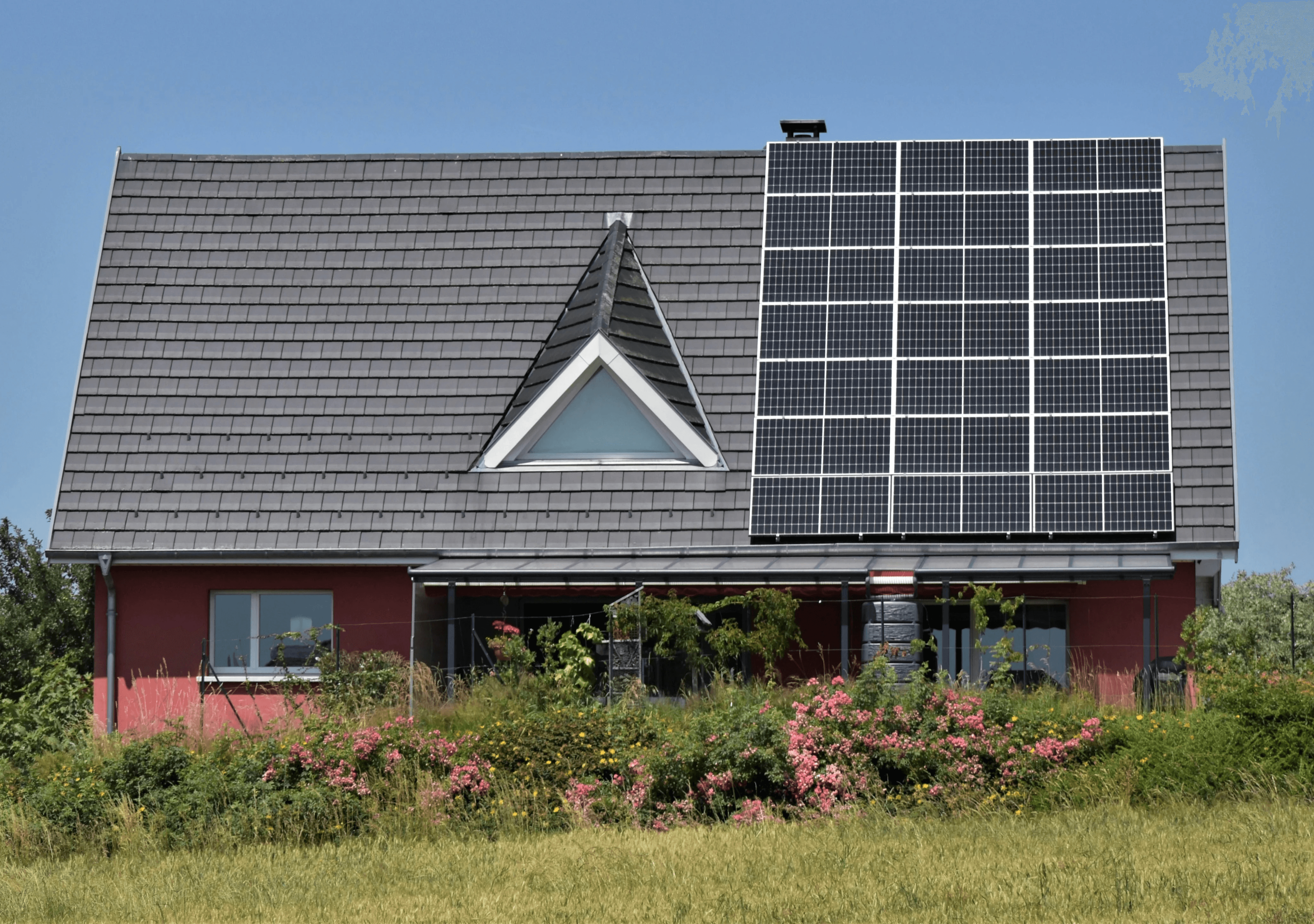 Maison moderne rouge avec toit gris équipé de panneaux photovoltaïques, entourée de jardin et de fleurs, captant l'énergie solaire pour une habitation durable.