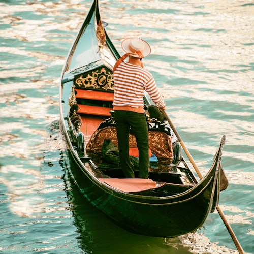 A gondolier in a striped shirt and hat rows a gondola through calm, greenish-blue water.