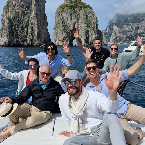 A group of smiling adults seated on a boat waving, with tall rocky cliffs and the ocean in the background.