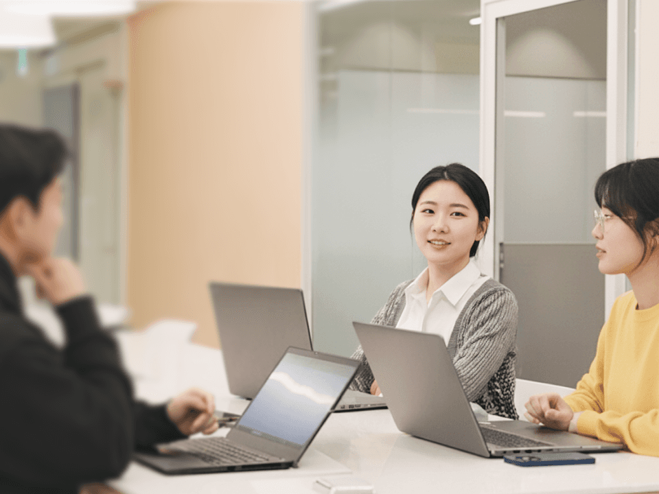 Three people sit at a table with laptops, engaging in a discussion in a modern office setting.