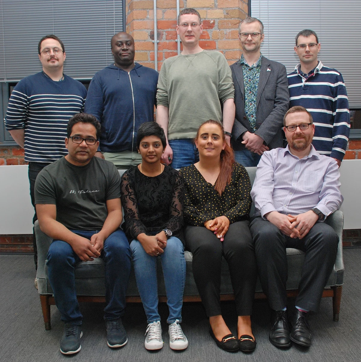 Ten people are posed for a group photo indoors, with some seated on a sofa and others standing behind. The setting features brick walls, blinds, and office-style lighting.