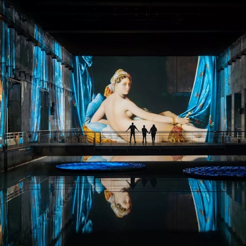 มีคนสามคนยืนอยู่หน้าภาพโปรเจกชั่นขนาดใหญ่ของภาพวาดผู้หญิงนอนในสไตล์คลาสสิก ซึ่งสะท้อนลงในน้ำด้านล่าง