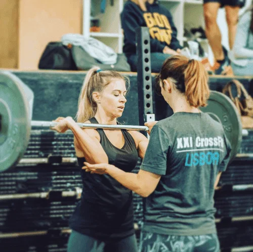 Uma atleta a segurar uma barra em posição de front rack outra a ajudar