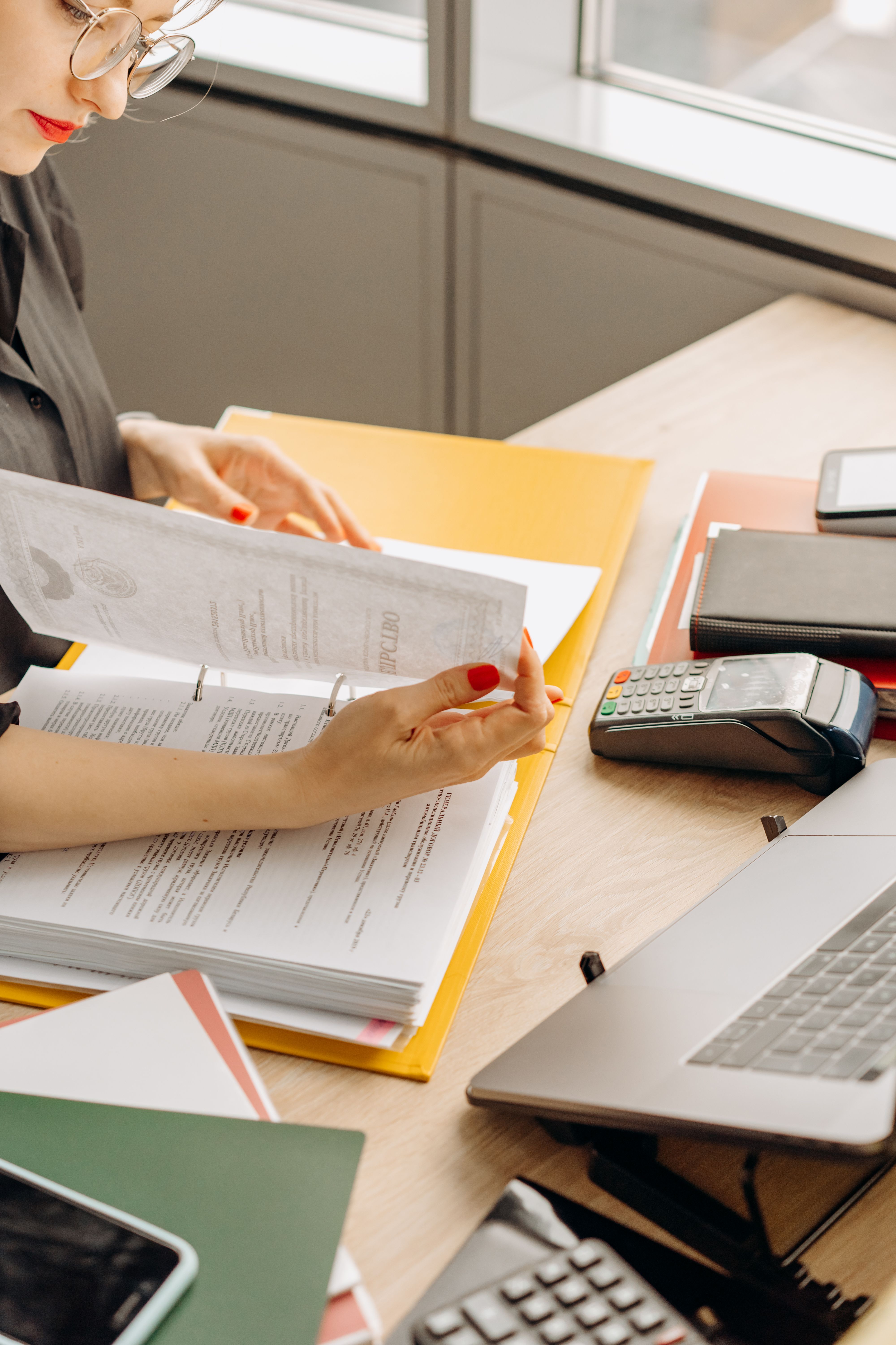 A person reviewing documents inside a bright yellow binder at a office desk equipped with a laptop and payment terminal.