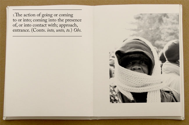 The image shows an open book resting on a light brown surface. The left page contains text defining the action of "going or coming to or into; coming into the presence of, or into contact with; approach, entrance." (Conts. into, unto, to.) Observed. The right page features a black and white photograph. The photo depicts a person wearing a hooded garment and a light-colored scarf or shawl, looking directly at the camera. The background is out of focus and shows indistinct shapes that might be other people.