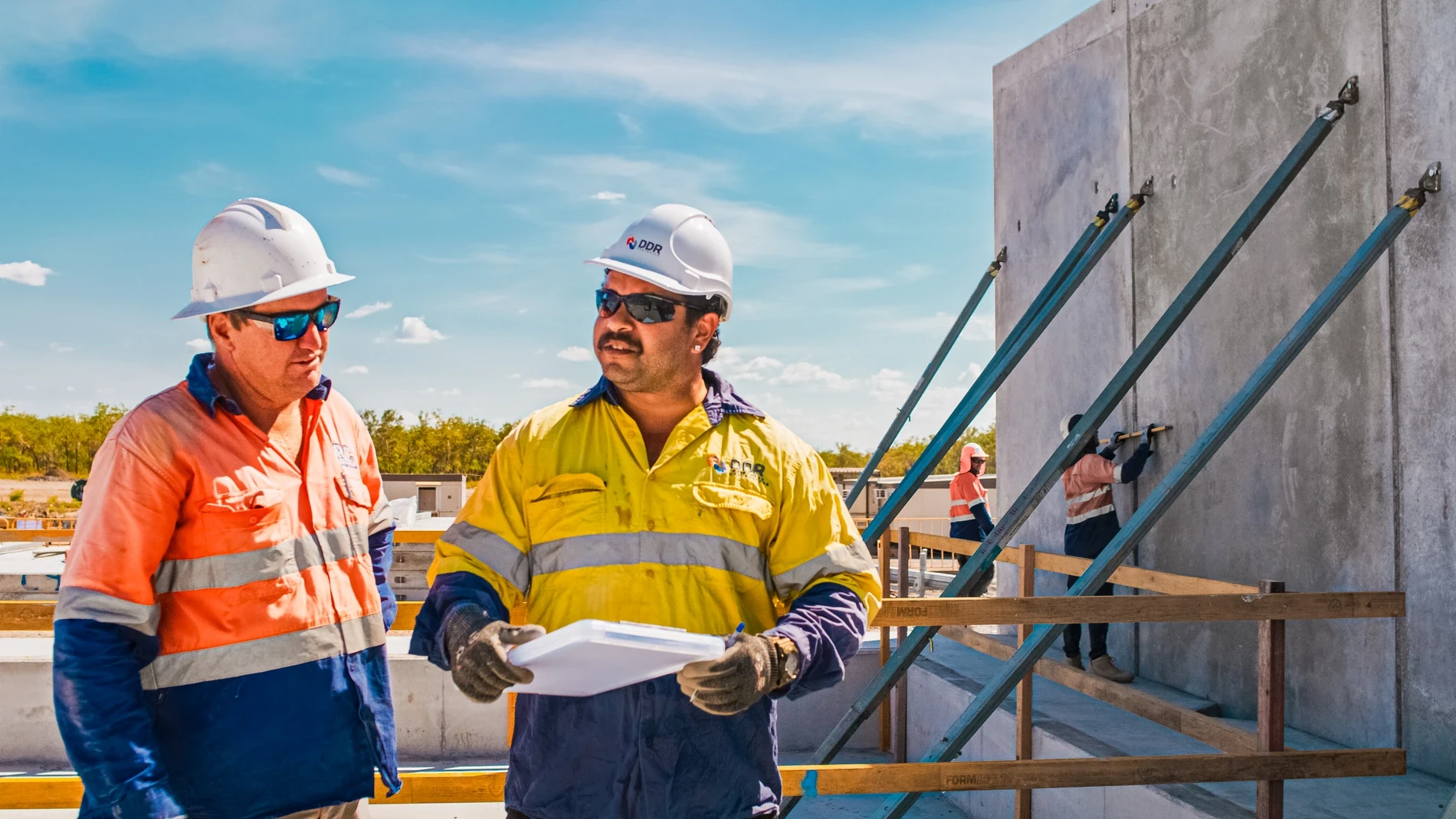Two workers in high-visibility clothing reading a drawing beside a propped concrete panel
