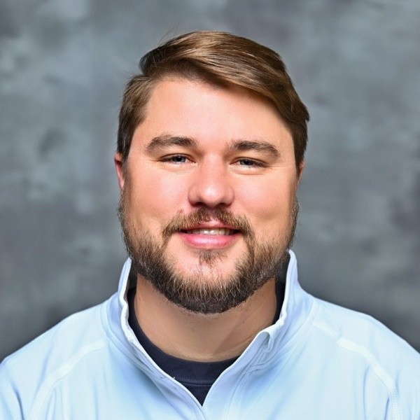 A smiling young woman with curly hair, wearing a purple shirt, stands in a bright indoor setting.