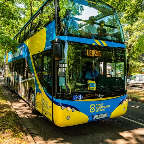 木々が並ぶ通りに停まっている、上部デッキに乗客がいる「ライプツィヒ」とラベルされた二階建て観光バス。