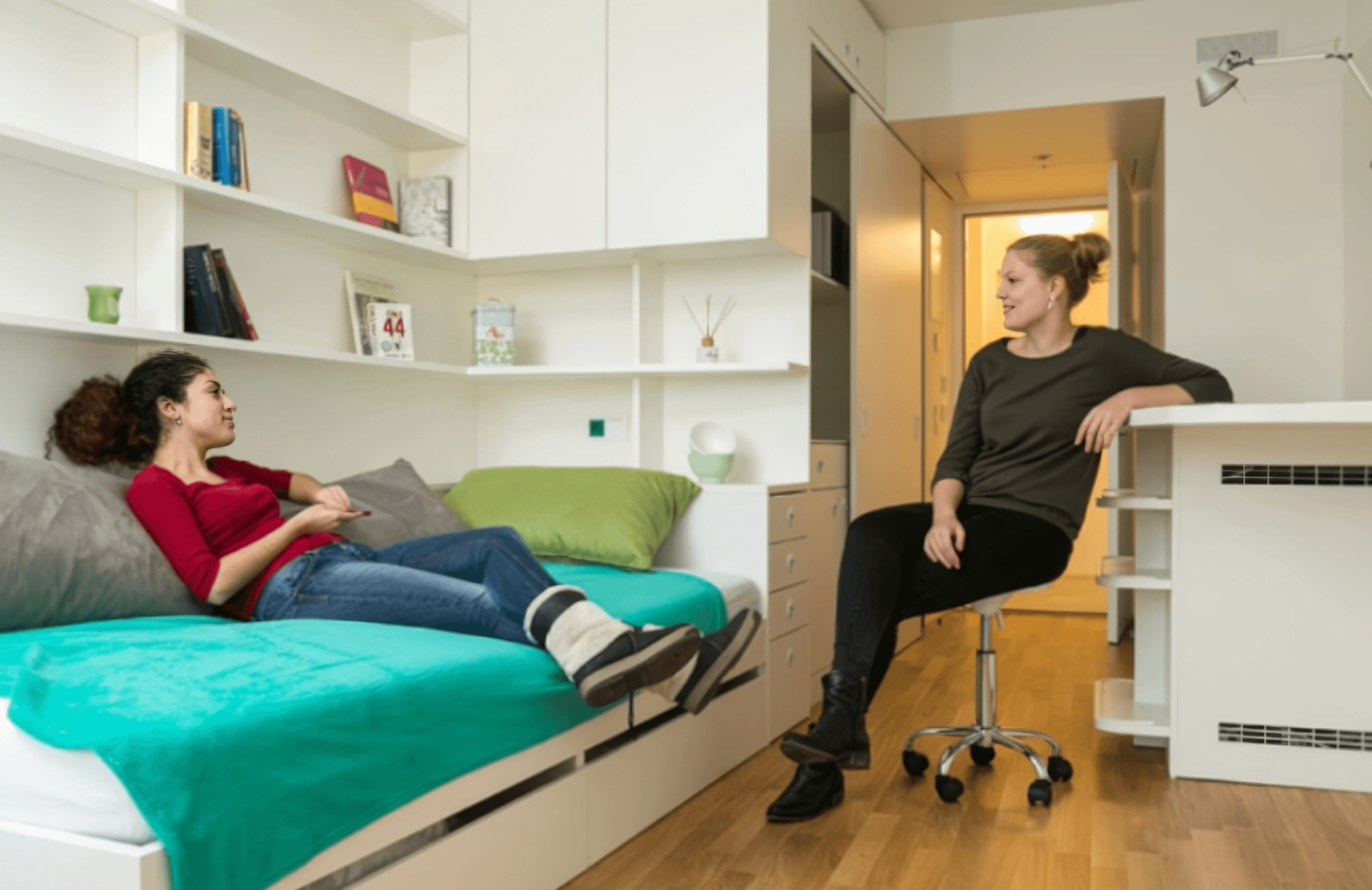 MW Interior - Zwei Studentinnen im gespräch in einem hellen weißen Studentenzimmer mit Bett, Regalwand, Schreibtisch und Holzparkett" Beschreibung: Lebendige Alltagsszene im Studentenzimmer: Integriertes Bett mit Stauraum darunter, wandseitiges Regalsystem, kompakter Schreibtisch und helles Parkett – ein Zimmer, das Wohnen, Schlafen und Lernen auf engem Raum komfortabel vereint.