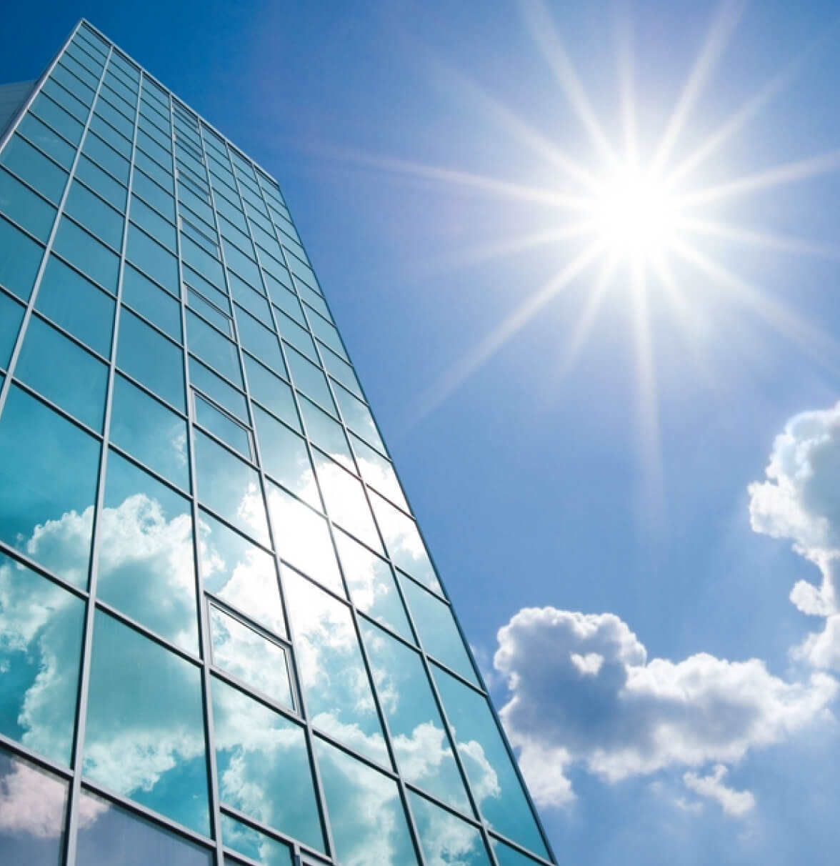 A tall glass building reaches towards a bright blue sky, reflecting sunlight and fluffy white clouds.