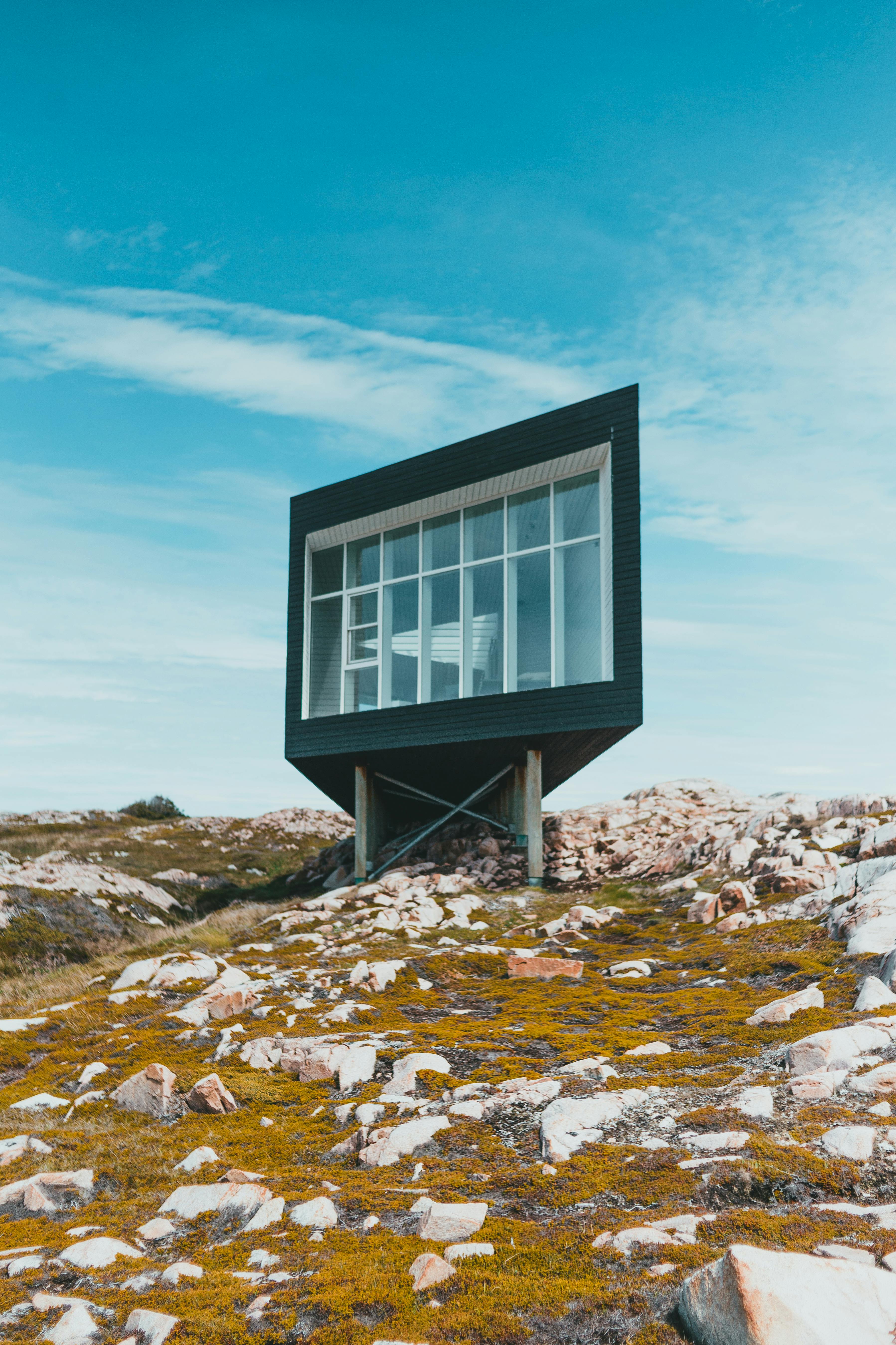 A modern, cube-shaped building with large glass windows is elevated on stilts over a rocky, moss-covered landscape under a clear blue sky.