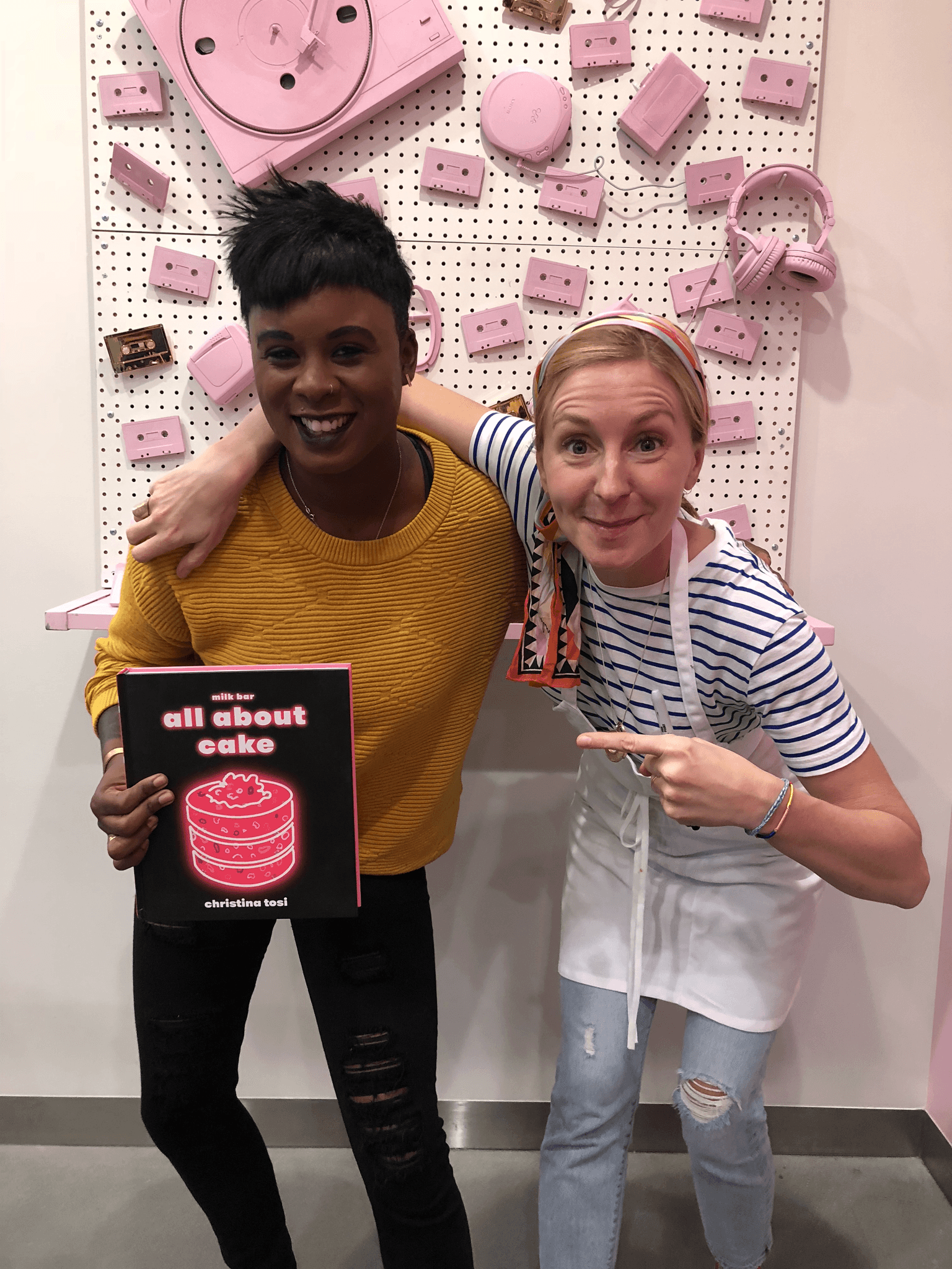 Ashleigh Warren and Christina Tosi smiling,  pose together, Ashleigh holding a book, in front of a pink wall adorned with decorations.