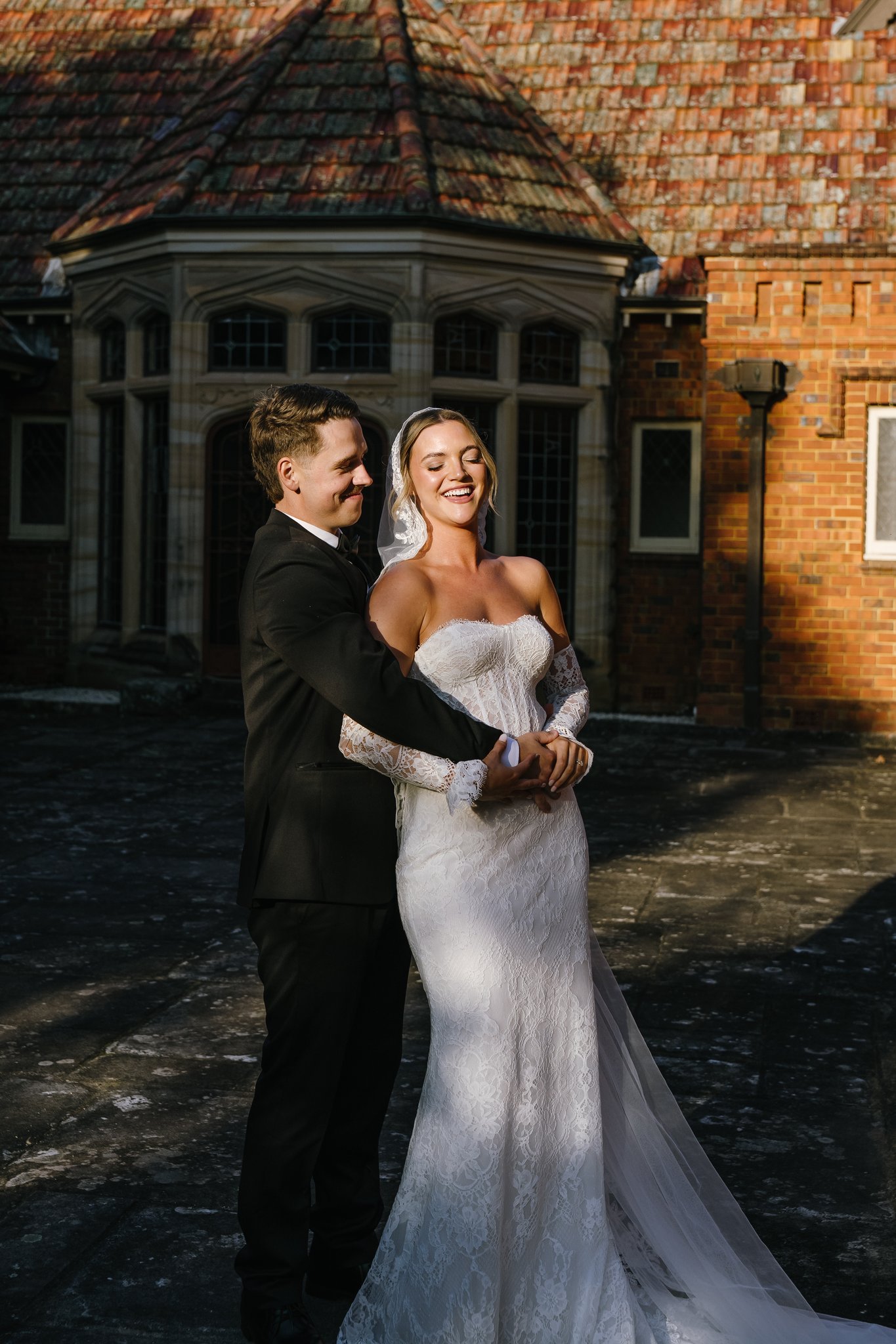 Groom embraces smiling bride in lace gown outside a brick mansion in warm sunlight.