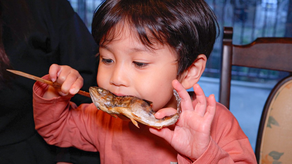 As part of the cultural and culinary experiences at our kids academy, a young student with a curious expression takes a bite of a whole grilled fish served on a traditional wooden skewer.