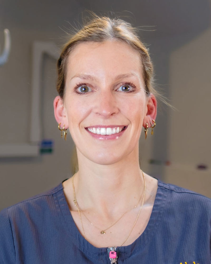 A portrait of Julia, Principal Dentist at Cricklade Dental Practice, smiling and wearing a dark grey scrub top with a pink zipper pull and yellow embroidered text that reads "JULIA" on the left and "Cricklade Dental Practice" on the right. She is standing in a dental surgery with a dental light and other equipment visible in the background.