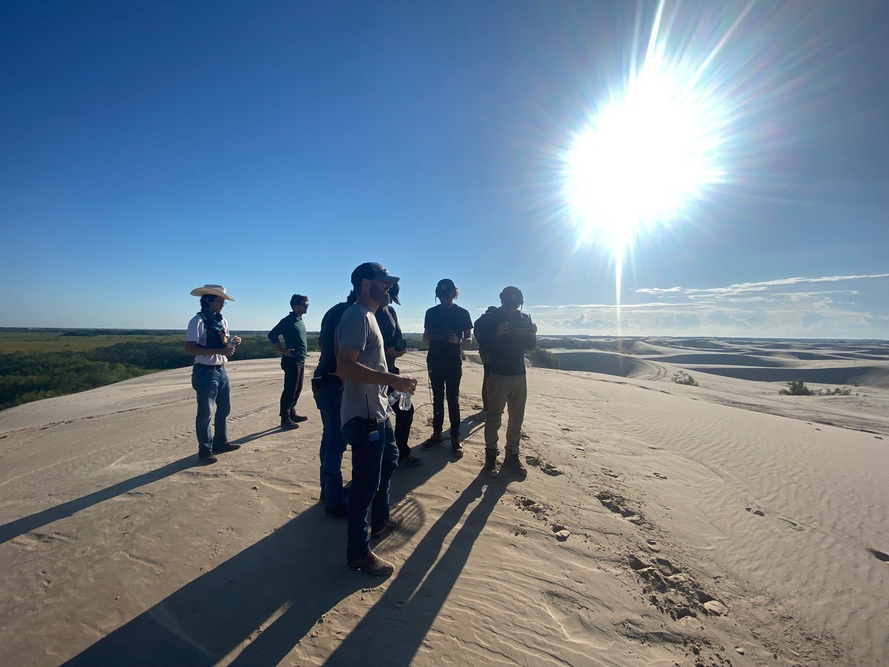 A live-action production crew stands atop sunlit sand dunes during a location scout or shoot setup. The group, dressed in casual work attire with gear in hand, surveys the vast landscape under a blazing sun. Long shadows stretch across the sand, hinting at early morning or late afternoon light, with camera prep or scene planning underway in the open desert environment.