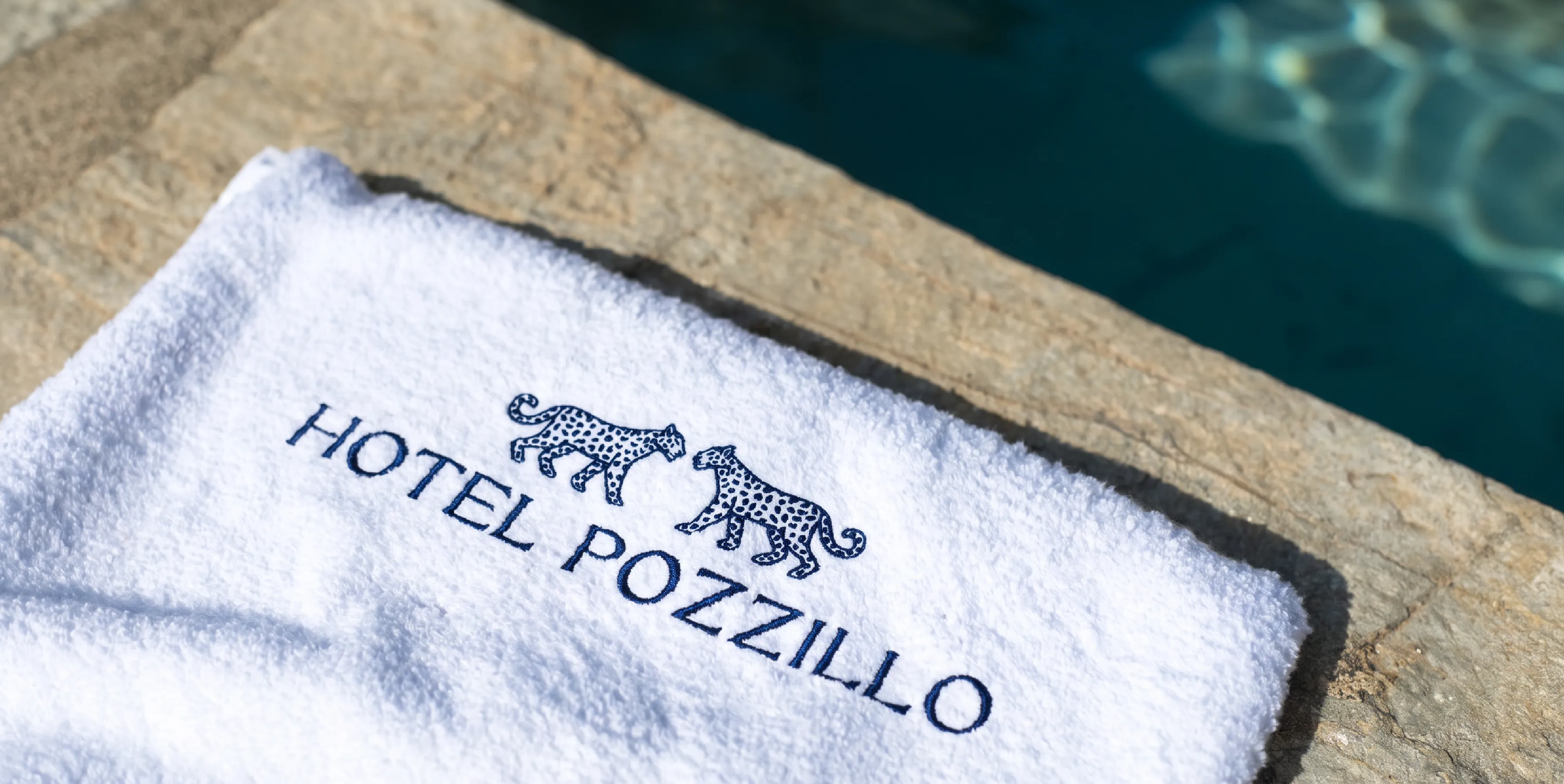 A close up of a white beach towel, placed poolside with the Hotel Pozzillo logo embroidered on