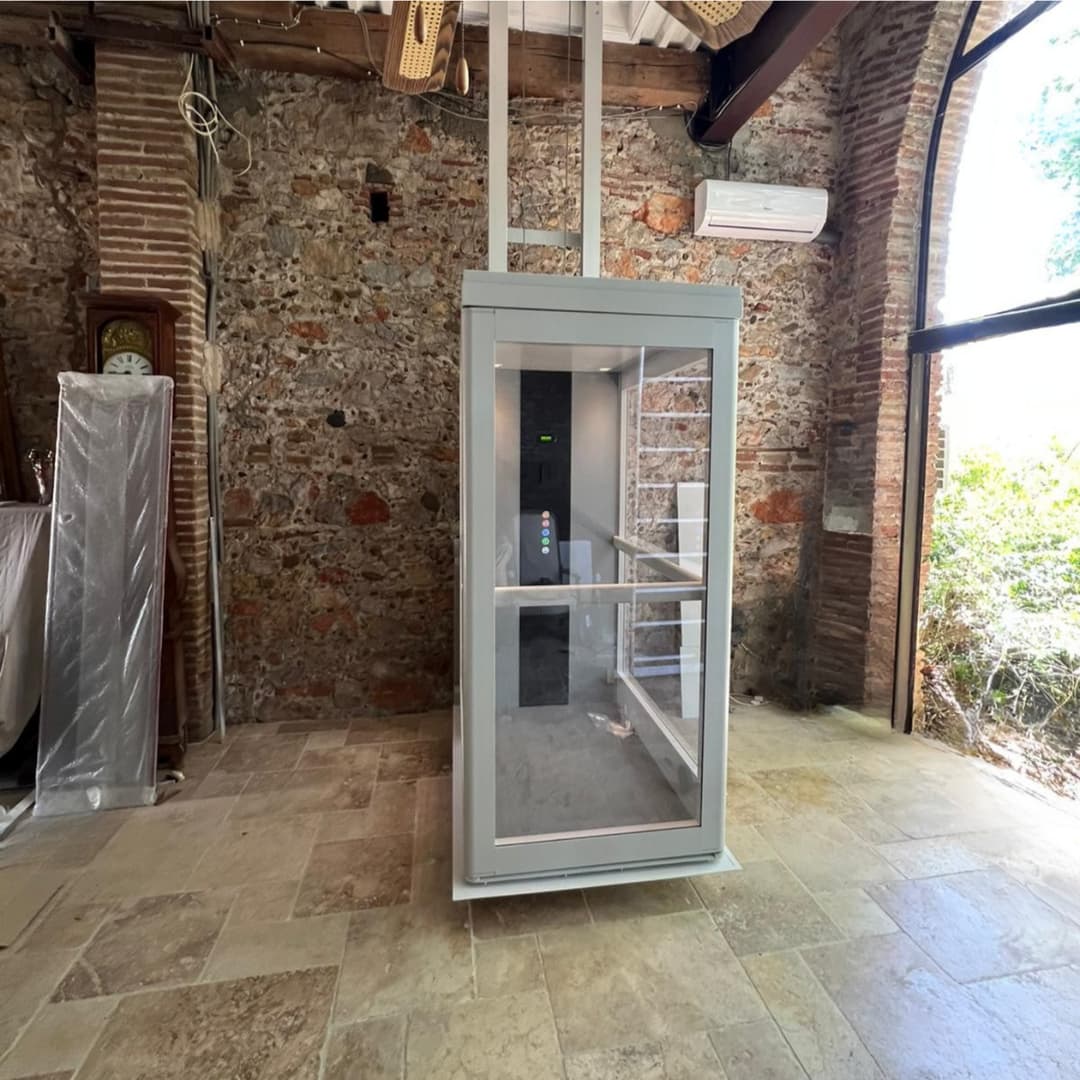 A glazed through-floor home lift with a grey frame and colour-coded floor controls, installed in a residential property with exposed stone walls and terracotta tile flooring.