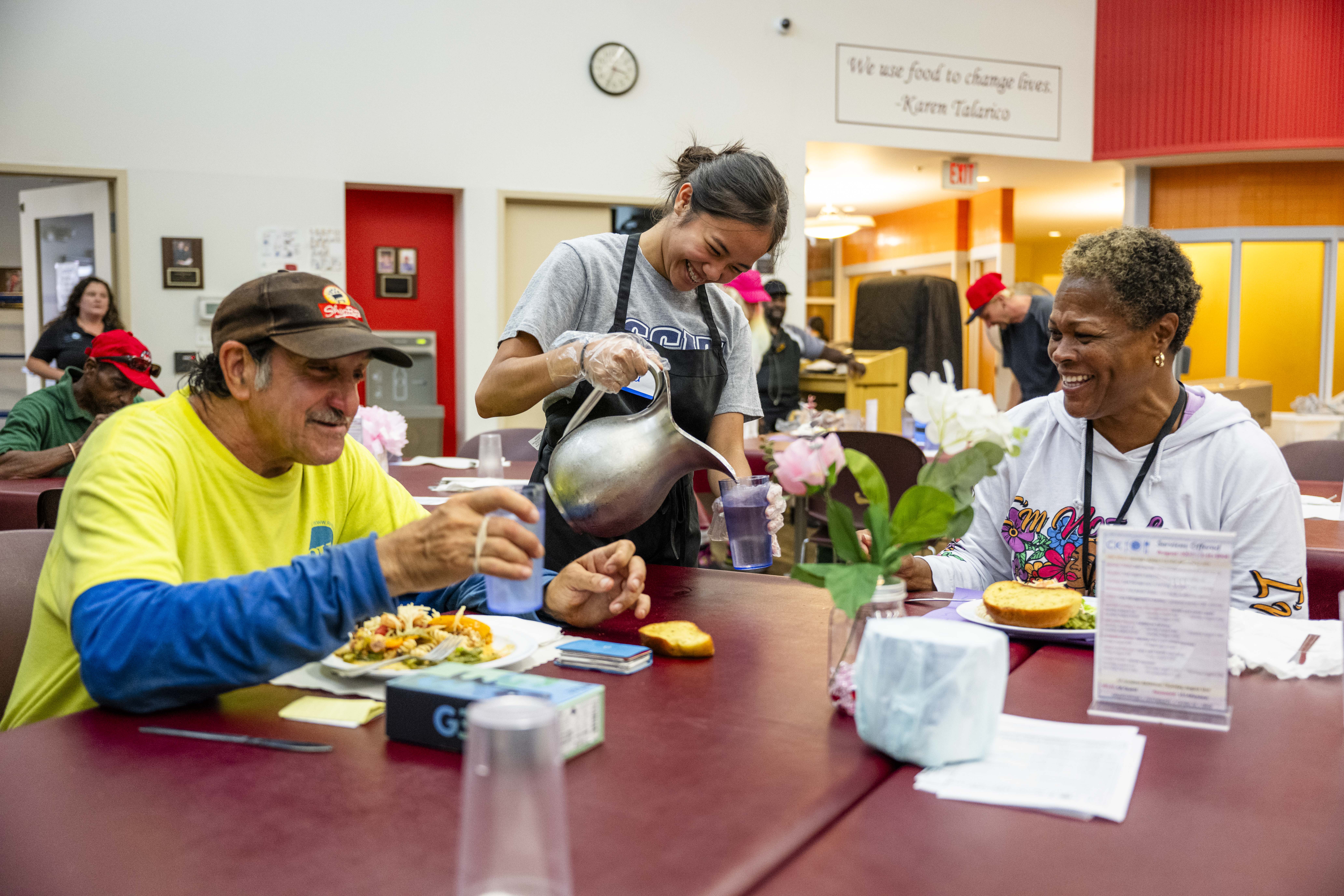Volunteer pours water in a glass for a guest in grantee Cathedral Kitchen's dining room. Two guests are eating nutritious meals and smiling as they sit at the table.