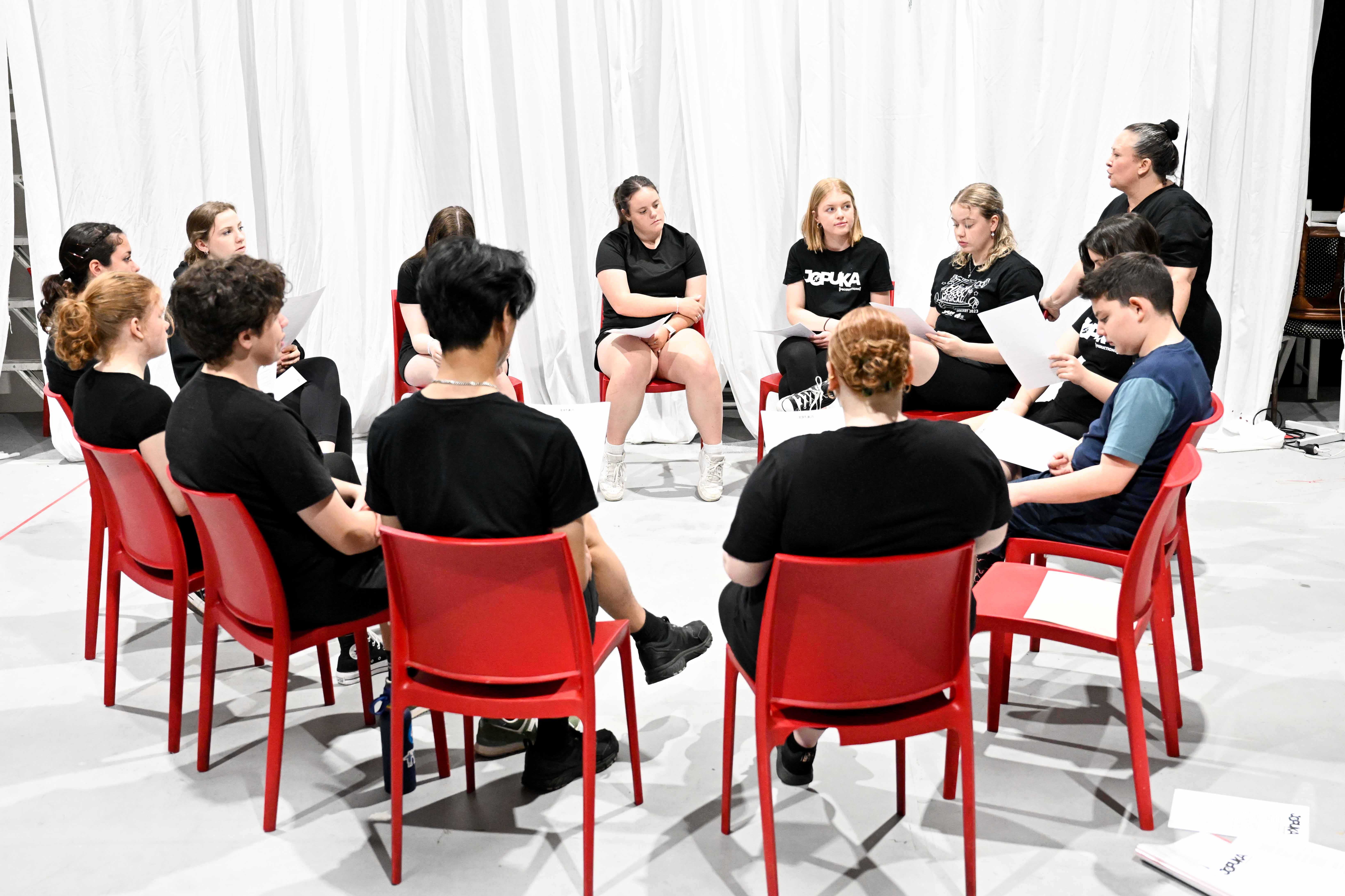 A group of young people sit in a circle on bright red chairs, holding sheets of paper during a workshop or rehearsal. All are dressed in casual black clothing, listening to an adult facilitator who stands beside them, speaking and guiding the discussion. The scene takes place indoors against a backdrop of long white curtains.
