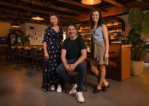 BarBucce founders Jacqueline, Michael, and Jennifer posing inside their warmly lit restaurant, with wooden beams and shelves of bottles in the background.