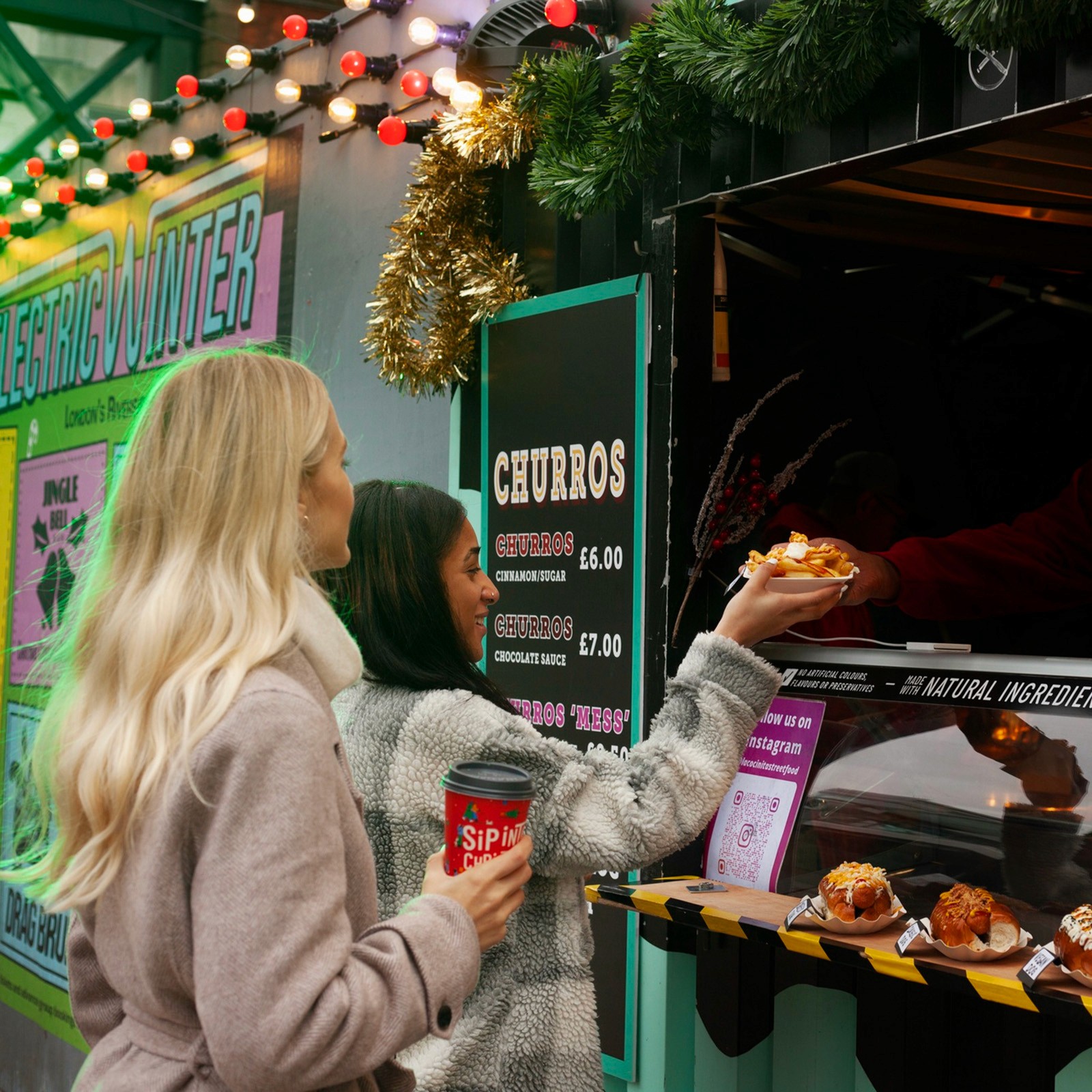 Two women at a festive outdoor winter market receiving a plate of food from a churro stand.