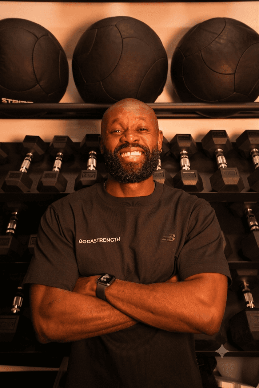 A medium close-up photograph of a bald, muscular man with a full beard, smiling warmly directly at the camera. He has his arms crossed over his chest, wearing a black t-shirt with text that reads 'GODASTRENGTH' on the left side and an 'NB' (New Balance) logo on the right side. He is also wearing a dark smartwatch on his left wrist and a silver ring on his right ring finger. He is standing in a brightly lit gym, in front of a black metal rack stocked with various dumbbells and larger black medicine balls above them. The background is composed of the multi-level dumbbell racks.