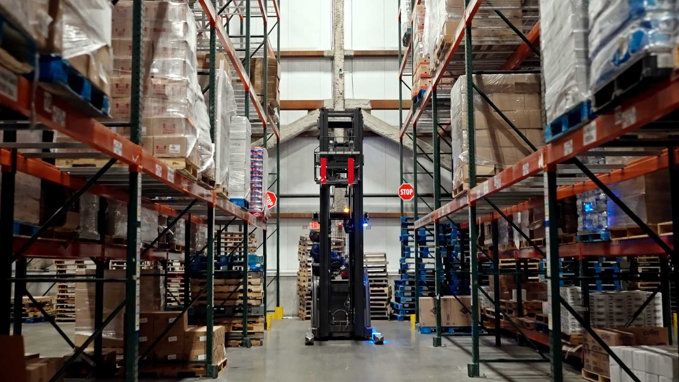 A forklift is centered in a wide warehouse aisle between tall pallet racks loaded with boxes and supplies.