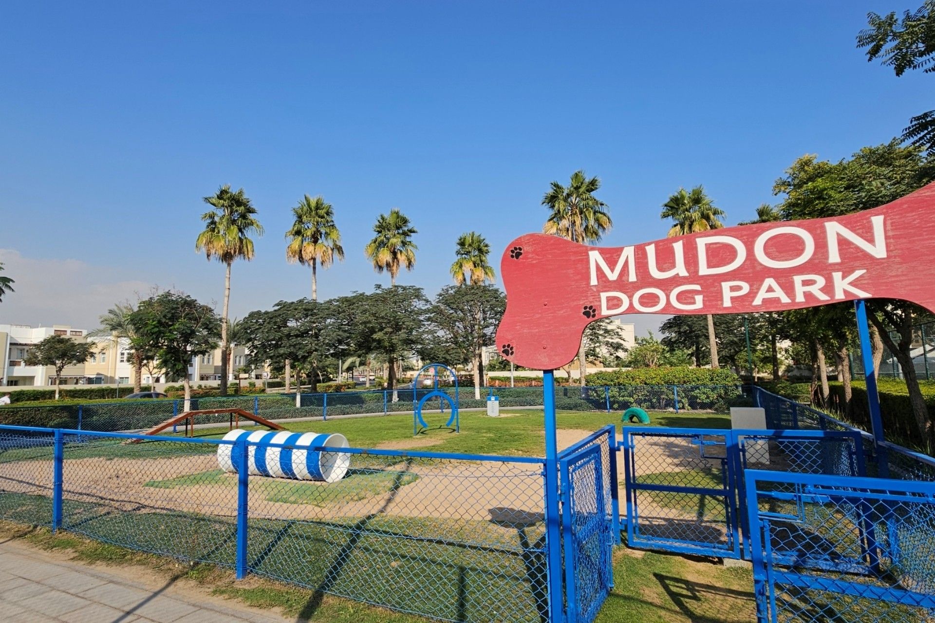 The Mudon Dog Park with a red sign, blue fencing, agility equipment, and greenery nearby.