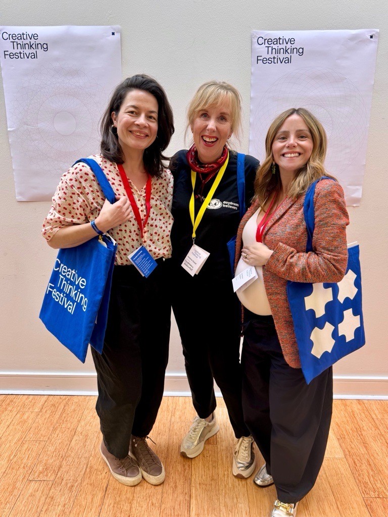 Three people stand side by side at an indoor event, each wearing a lanyard and holding branded tote bags. Posters reading “Creative Thinking Festival” are displayed on the wall behind them, and the group is standing on a wooden floor against a light‑coloured background.