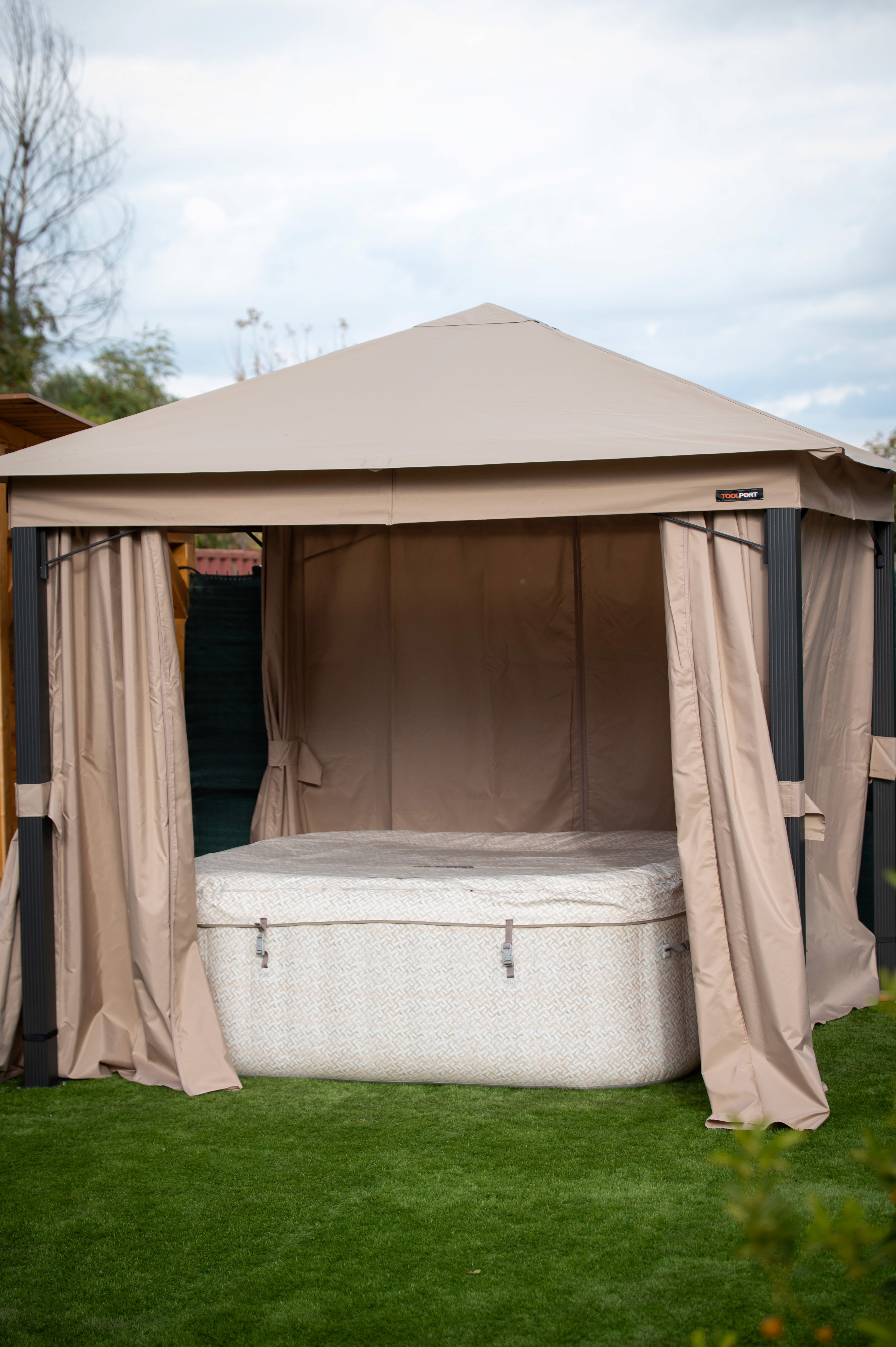 Egy bézs színű baldachinos sátor nyitott függönyökkel áll a zöld gyepen, alatta egy négyzet alakú, pöttyös mintájú hordozható pezsgőfürdővel, felhős égbolt hátterében.