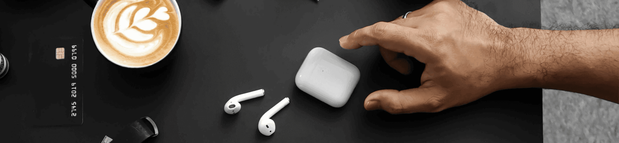 A hand reaching across a black table, several items are visible, coffee, airpods, watch and a bank card