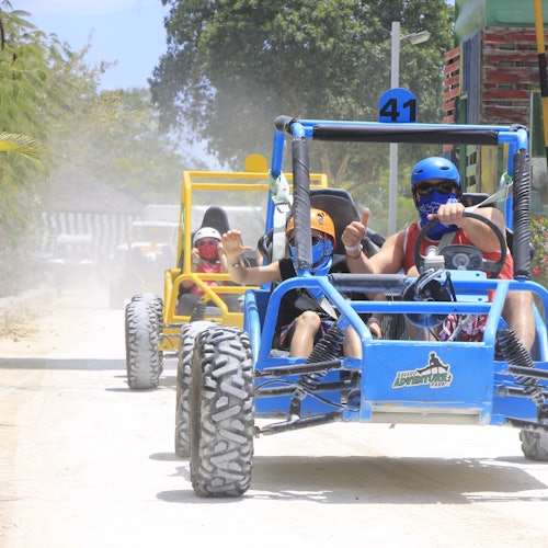 People wearing helmets and masks drive dune buggies on a dirt path, kicking up dust, with trees and a building in the background.