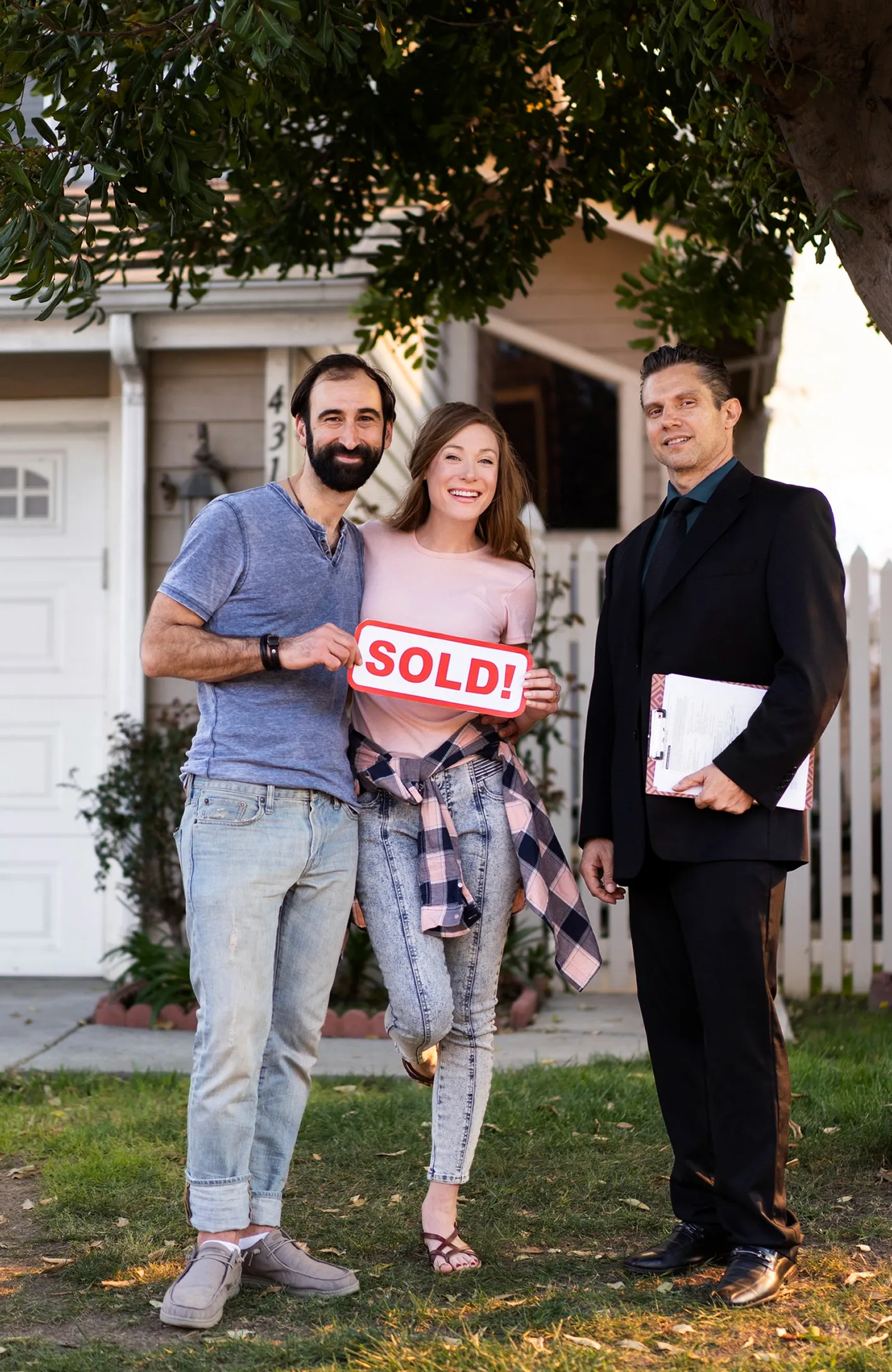 A couple stands outside their home, smiling, with a man in a suit holding paperwork nearby.