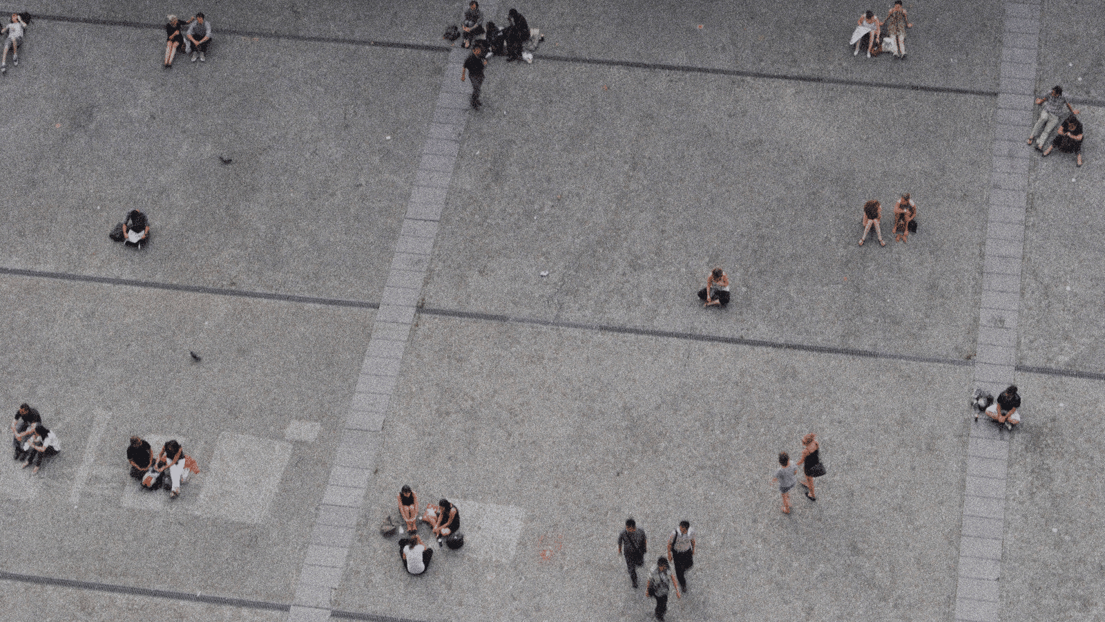 Aerial view of people scattered across a city plaza, seated or walking in small groups on a grid-like pavement.