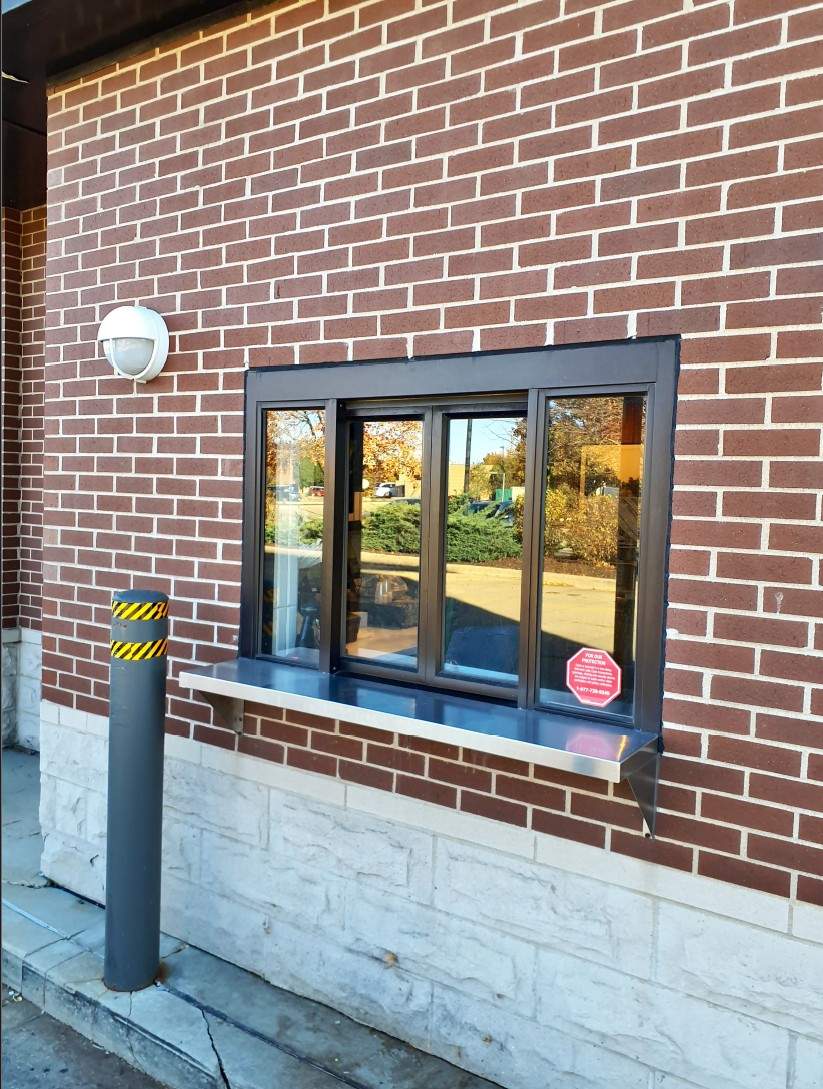 Exterior drive-thru window framed in dark metal and brick, featuring stainless trim and protective bollard.