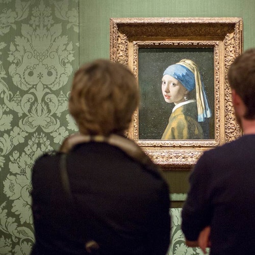 Dos personas observan la pintura "La joven de la perla" en un museo con papel tapiz verde con patrones.