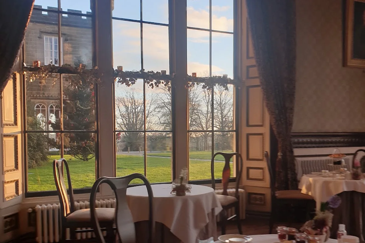 A country house breakfast room: tall arched windows looking out over lawns, round tables laid with white linen, in low winter light.