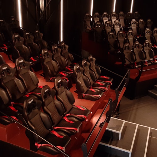 Monitasoinen istuma-alue, jossa on riveittäin mustia ja punaisia pelituoleja tai elokuvateatterituoleja, varustettuna käsinojilla ja turvavöillä.