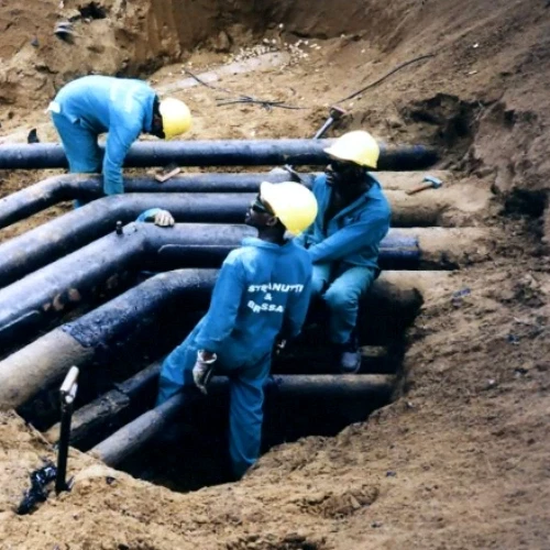 Arbeiter in blauen Overalls und gelben Helmen installieren schwarze Rohre in einem Graben. Baustelle.